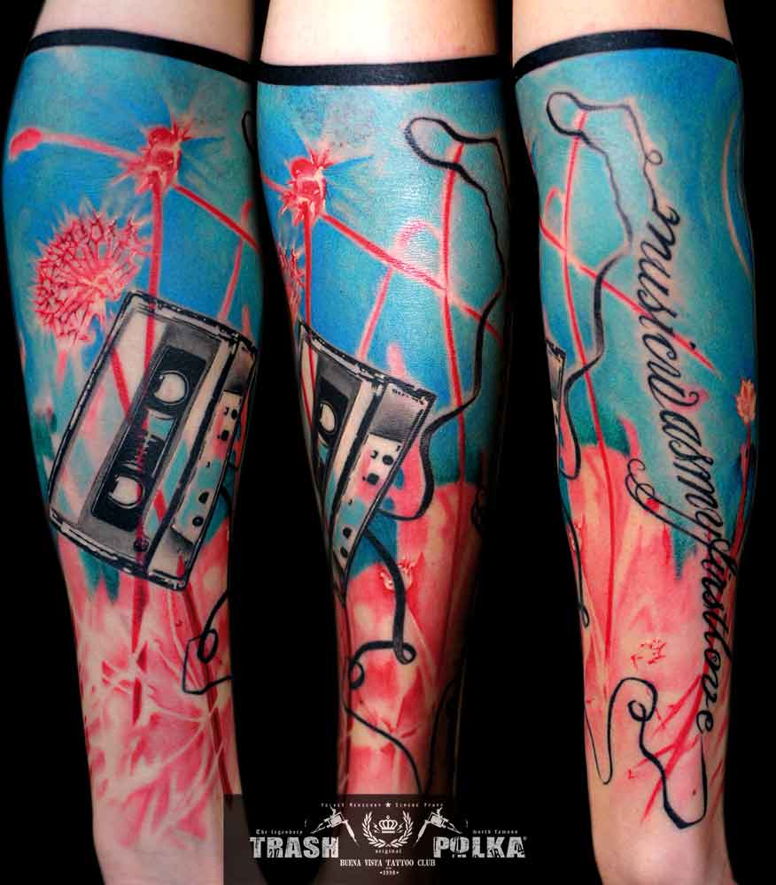 trash polka tattoo on the forearm very colorful a cassette with a hanging tape ending in writing and light blue clouds and pink dandelions
